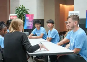 A group of middle school students and two adults sit around a table talking.